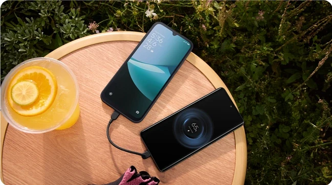 Dwa telefony Redmi 15C leżące na okrągłym stoliku w plenerze, jeden w trakcie ładowania