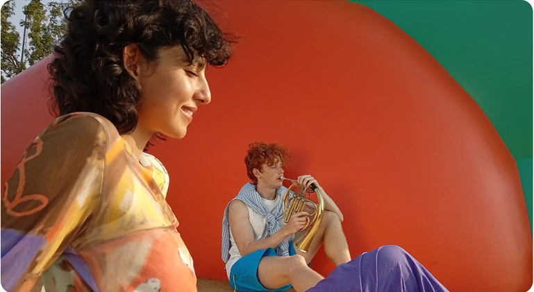 Dwoje młodych osób w kolorowych strojach na tle dużej pomarańczowej struktury, zdjęcie wykonane Redmi 15C